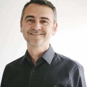 A man in a black shirt smiling in front of a white background.